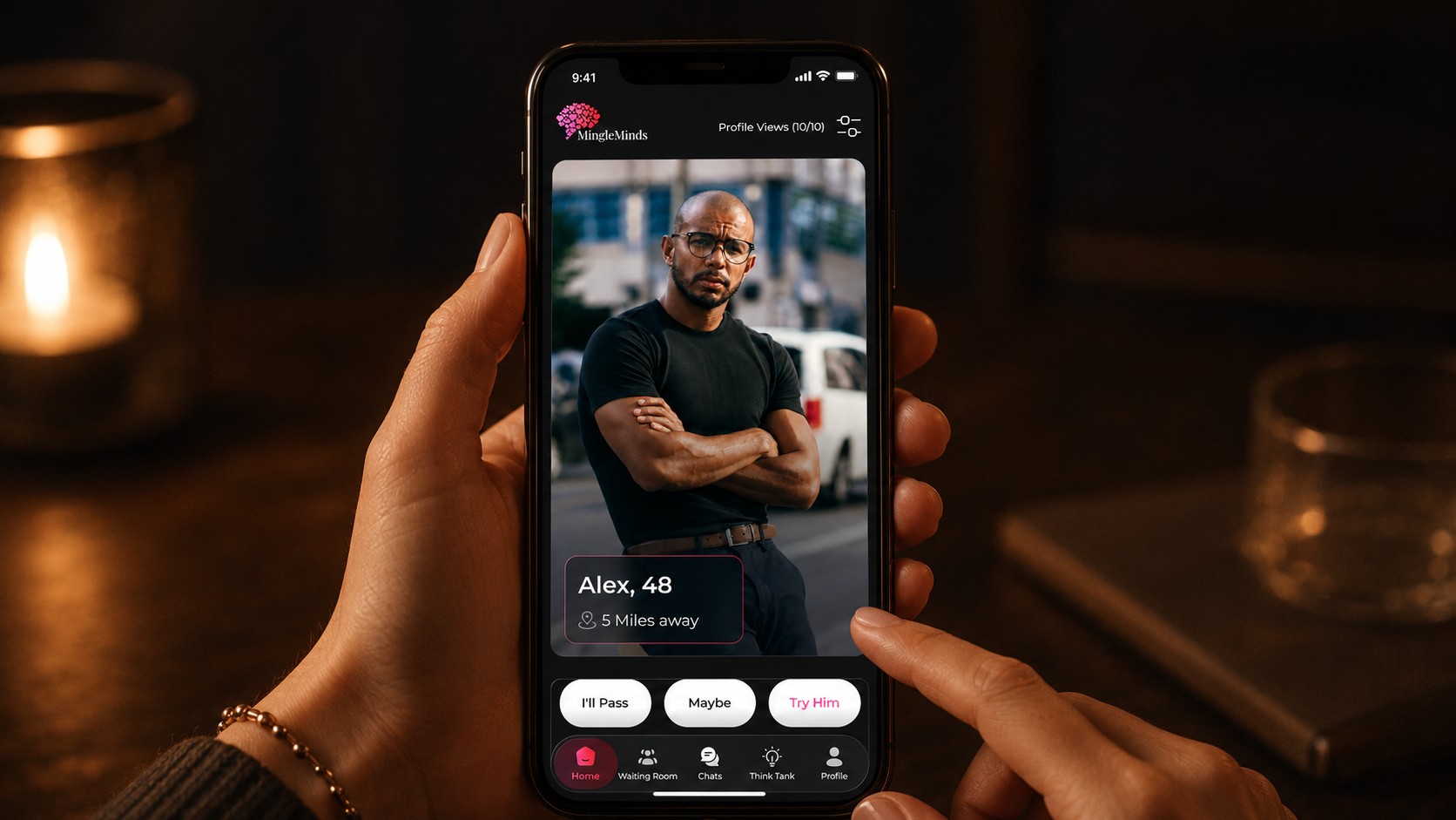 A woman's hands hold her phone upright, browsing a MingleMinds profile — Alex, 48 — with I'll Pass, Maybe and Try Him beneath it, on a quiet table lit by a candle.